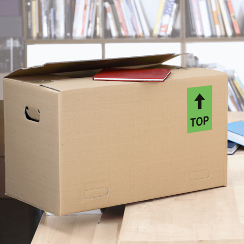 Kartonbox mit einem roten Heft auf dem Deckel, beschriftet mit einem grünen "TOP"-Schild. Im Hintergrund sind Bücherregale sichtbar.