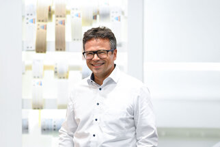 A man wearing glasses and a shirt stands smiling in front of a bright, modern backdrop featuring various materials.
