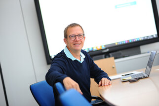 A man with glasses is sitting at a table in a conference room, smiling, with a laptop in front of him.
