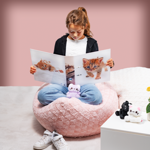 A girl is sitting in a large, soft bean bag and looking at a book filled with pictures of kittens. Plush toys are scattered around her.