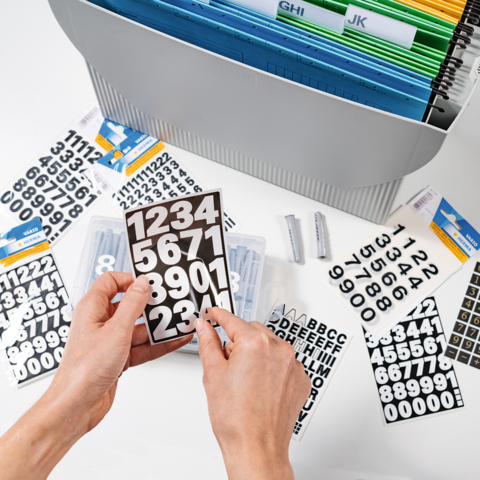 Hands hold a black board with white numbers, surrounded by various numbered and alphabetical stickers in an organizer.