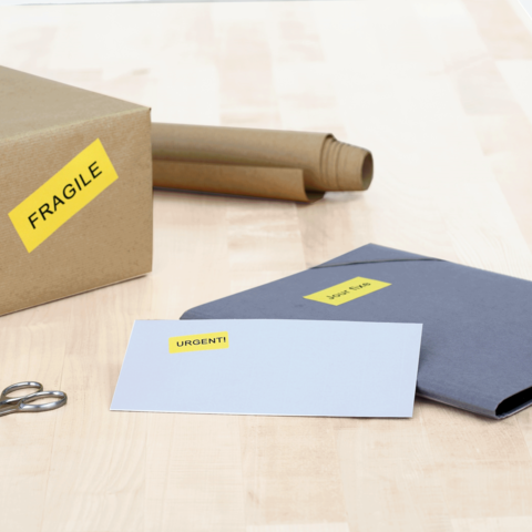 Ein Karton mit dem Aufkleber "FRAGILE", ein graues Kuvert mit "DRINGEND!" und eine Schere auf einem Holztisch.