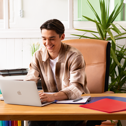 Ein junger Mann sitzt an einem Schreibtisch mit einem Laptop, umgeben von Schreibmaterialien und Pflanzen, und arbeitet konzentriert.