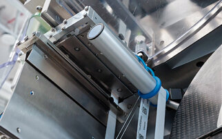 Close-up of a mechanical part featuring a silver cylinder and blue rubber bands, mounted on a metallic structure.