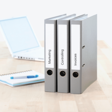 Drei graue Ordner mit Beschriftungen "Marketing", "Controlling" und "Rechnungen" stehen neben einem Laptop und einem Notizblock.