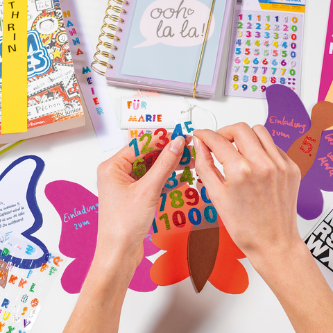 Hands hold colorful butterfly-shaped invitations, surrounded by a notebook and stickers.