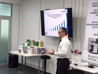 A man is presenting in front of a screen displaying a chart about digital printing machines. Various printing materials are visible in the background.
