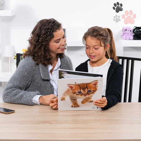 A woman and a girl are sitting at a table, looking at a picture book about cats together.