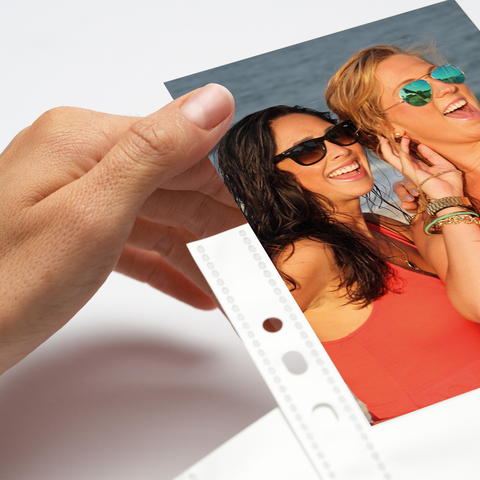 A hand holds a photo of two women wearing sunglasses and smiling, with a water background.