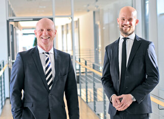 Two men in suits are standing in a modern office hallway, smiling. The man on the left is wearing a striped shirt, while the one on the right has a watch.