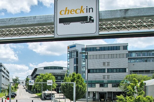 A modern building complex with a pedestrian bridge, featuring traffic signs and a 'Check-in' sign, surrounded by trees and a blue sky.