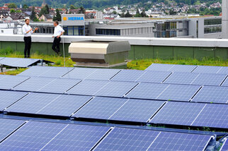 Two people in suits are standing on a roof with solar panels, with buildings and a cityscape visible in the background.