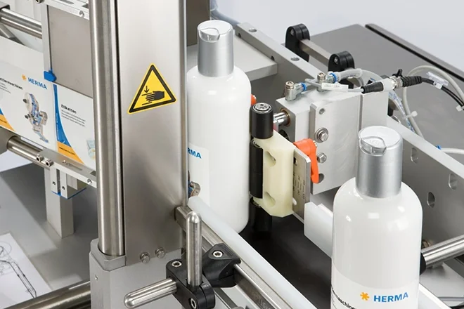 A bottling machine with white bottles on a conveyor belt, equipped with various technical components and warning signs.