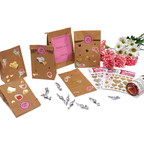 Geschenktüten aus Kraftpapier mit Aufklebern, Süßigkeiten, Herzmotiven und einer Flasche Roséwein, umgeben von Blumen.