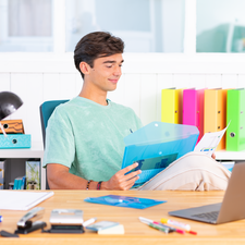 Ein junger Mann sitzt an einem Schreibtisch, hält eine transparente Mappe und lächelt. Im Hintergrund sind Ordner und ein Laptop sichtbar.