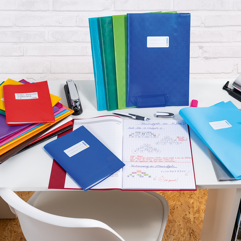 A desk with colorful folders, notebooks, and writing supplies. In the background, stacks of notebooks and a stapler are visible.