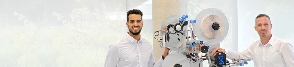 Two men in white shirts are standing next to a technical machine with various components and controls.