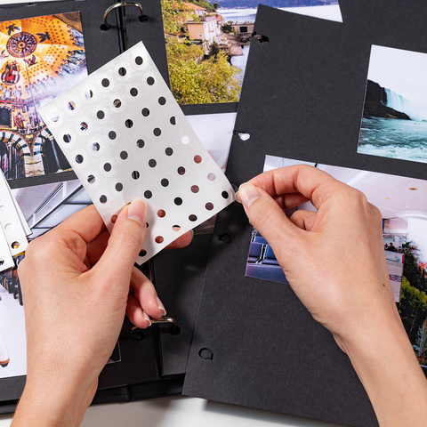 A person is holding a transparent sheet with dots in one hand while flipping through a photo book filled with various images.