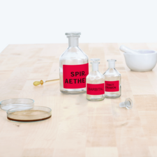 Flasche mit rotem Etikett "Spir. Aether", zwei kleinere Flaschen mit "Barbital" und "Tinct. Tramon", sowie ein Mörser und Petrischalen auf einem Tisch.