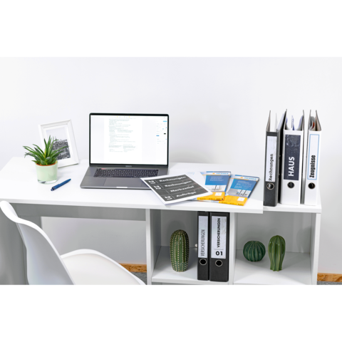 Schreibtisch mit Laptop, Aktenordnern und Dokumenten. Eine Pflanze und ein Bilderrahmen sind ebenfalls sichtbar.