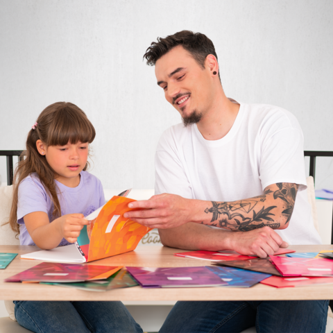 Ein Mann und ein Mädchen sitzen am Tisch und arbeiten gemeinsam an bunten Bastelmaterialien. Der Mann zeigt dem Mädchen etwas auf einem Blatt.