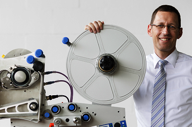 A man in a shirt and tie is holding a large disc and standing next to technical equipment with hoses and connections.