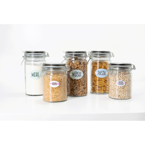 Fünf Glasbehälter mit Etiketten für Mehl, Reis, Müsli, Pasta und Haferflocken, auf einer weißen Oberfläche angeordnet.