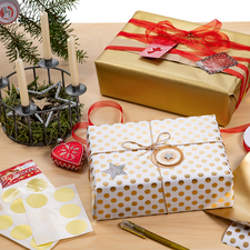 Geschenkpäckchen in gold und weiß mit Punkten, umgeben von Geschenkpapier, Dekorationen und einer Kerzenhalterung mit Tannenzweigen.
