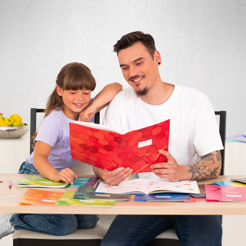 Ein Vater und seine Tochter sitzen an einem Tisch, während sie gemeinsam in einem bunten Buch blättern. Um sie herum liegen verschiedene Hefte und Materialien.