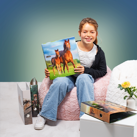 A girl is sitting in an armchair, holding a picture of two horses in her hand. Next to her are some binders and a plant.