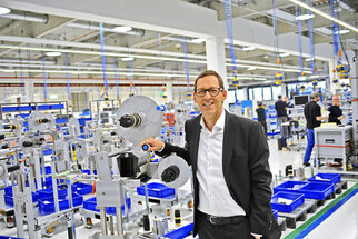 A man in a suit stands in a modern production hall holding a device. In the background, workstations and blue containers are visible.