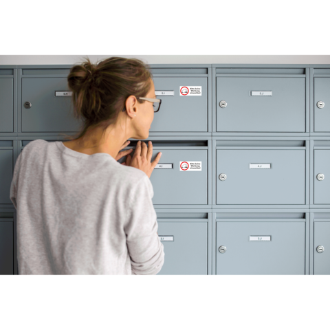 Eine Frau mit Brille schaut in einen grauen Briefkasten und überprüft den Inhalt. Die Briefkästen sind nummeriert und haben Aufkleber.
