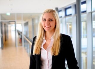 Young woman with long blonde hair, smiling in a modern office building while wearing a black blazer and a white shirt.
