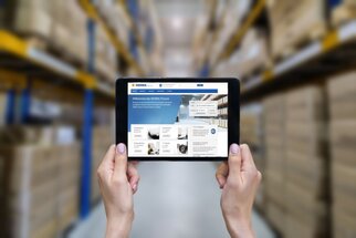 With a tablet in hand, it displays a website with information about inventory management, while shelves filled with boxes are visible in the background.