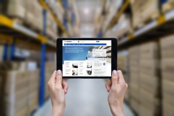 With a tablet in hand, it displays a website with information about inventory management, while shelves filled with boxes are visible in the background.