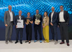 A group of six people stands on a stage, holding awards and flowers. In the background, a digital pattern is visible.