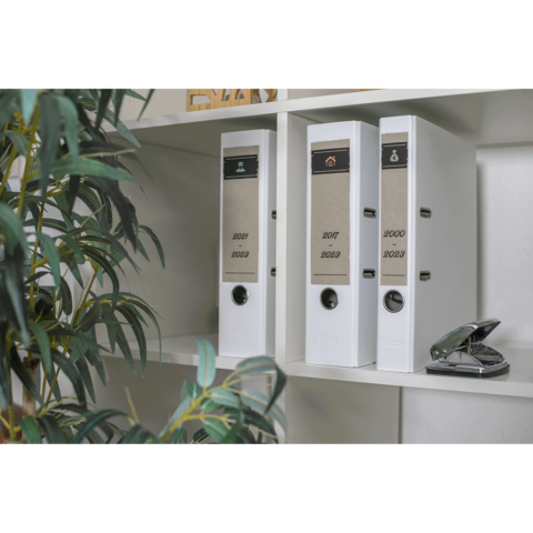 Drei weiße Ordner auf einem Regal, beschriftet mit den Jahren 2020, 2021 und 2022. Ein Heftgerät steht daneben, umgeben von einer Pflanze.