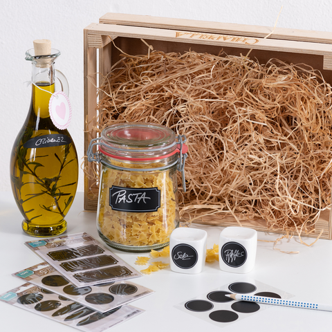 A glass of pasta, a bottle of olive oil, two spice containers, and stickers are placed in a wooden box filled with hay.