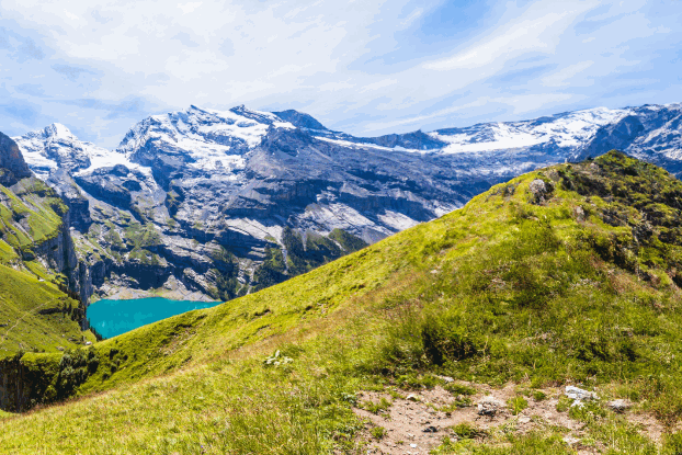 Panoramic view of green hills and snow-capped mountains, surrounded by clear water and a blue sky.