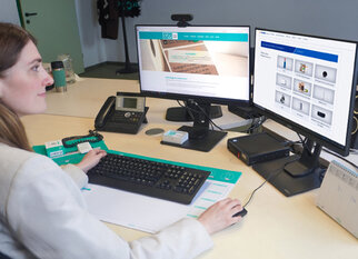 A woman is sitting at a desk, working on two monitors, with a phone and office supplies nearby.