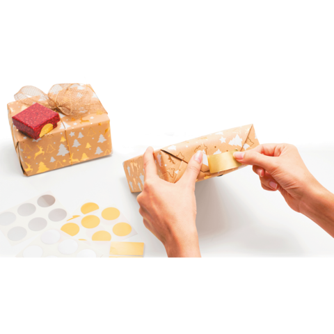 Eine Person wickelt ein Geschenk in braunes Papier mit goldenen und silbernen Motiven und verwendet Klebeband. Im Hintergrund sind weitere Geschenke und Aufkleber zu sehen.