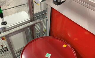 Red container with a label and a yellow lid, placed in an industrial setting with machines in the background.