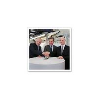 Three men in suits stand around a round table, placing their hands on a central object.