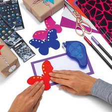 Hands are creating colorful butterflies out of paper, surrounded by crafting materials like gifts, stickers, and markers.