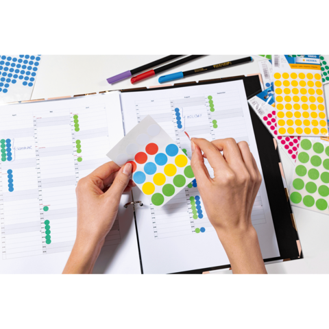 Hände halten bunte Aufkleber über einem geöffneten Planer mit Wochenübersicht. Stifte liegen daneben.