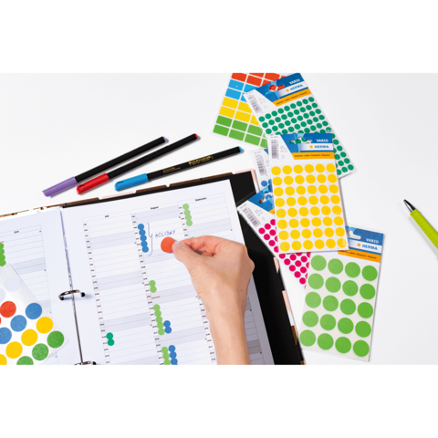 Ein Hand greift nach einem roten Aufkleber, während ein Planer mit farbigen Punkten und Stiften auf einem Tisch liegt.