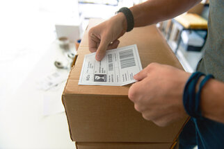 A person attaches a shipping label to a box while working at a table with more packages and office supplies.