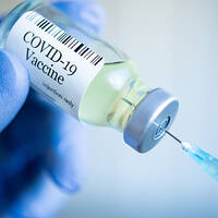 A hand in blue gloves holds a COVID-19 vaccine vial, while a syringe is ready to draw out the liquid.
