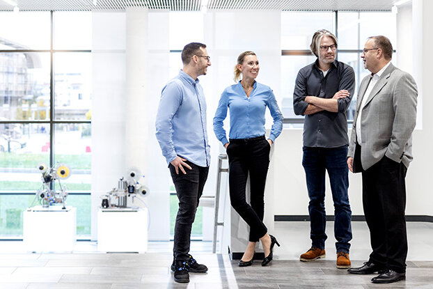 Four people are talking to each other in a modern office, while in the background, an industrial building and a sprawling facility can be seen.