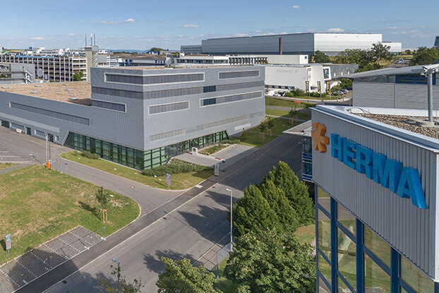 Four people are talking to each other in a modern office, while in the background, an industrial building and a sprawling facility can be seen.